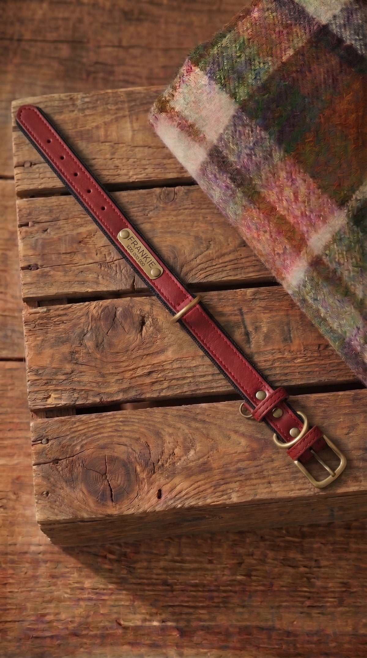 A close-up of a brown leather belt strap with brass hardware and a gold-toned buckle, laid diagonally on a rustic wooden crate next to a folded multicolor plaid textile.