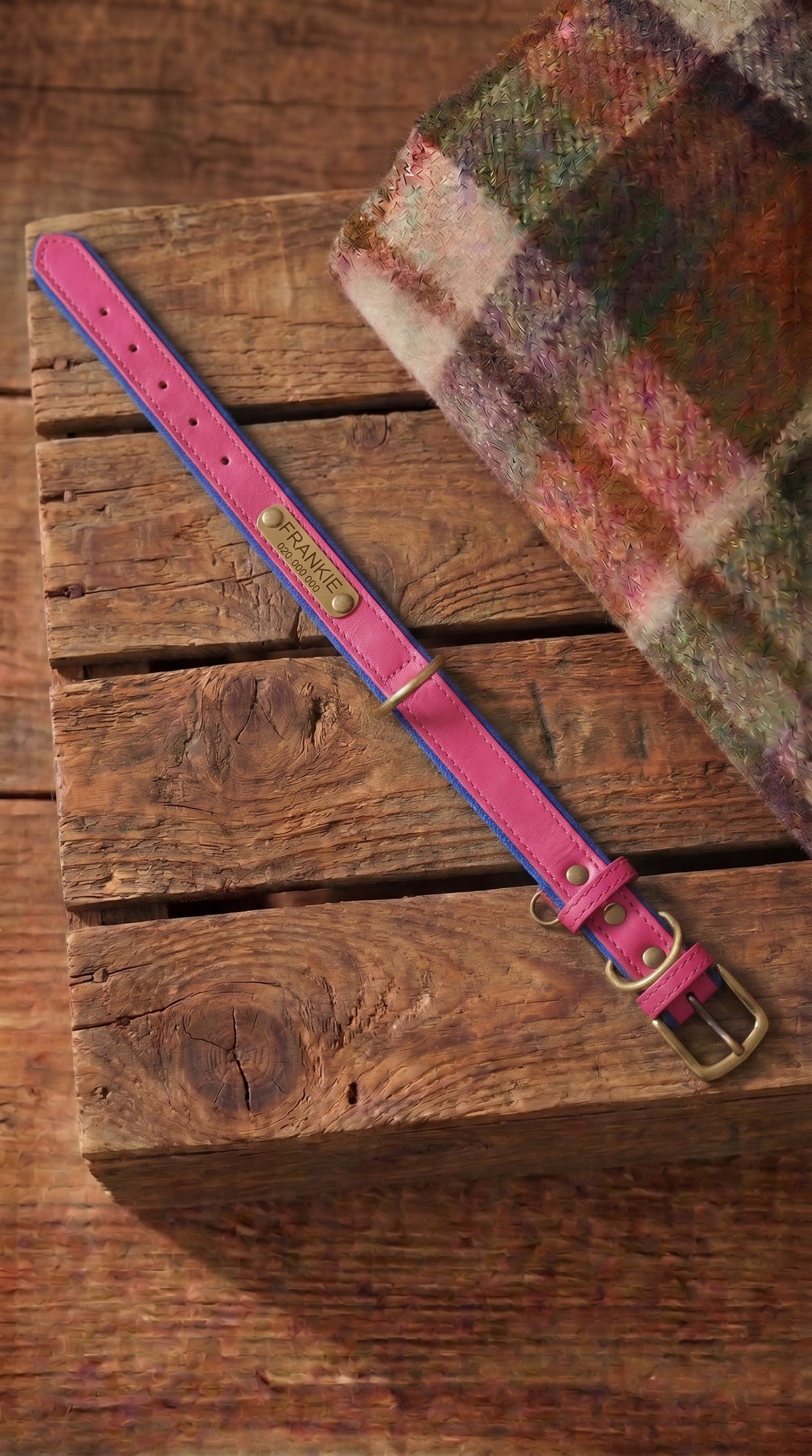 A pink leather dog collar with blue stitching and a brass buckle, laid diagonally on a rustic wooden crate next to a plaid wool blanket.