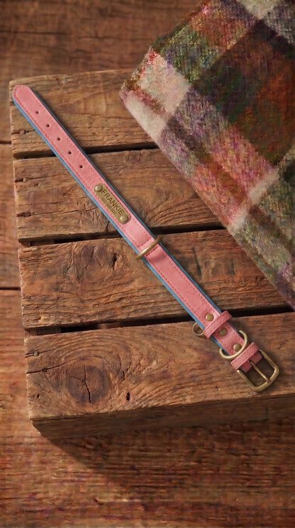 A pink leather belt with blue edge stitching and brass hardware, laid diagonally on a rustic wooden crate next to a plaid wool blanket.