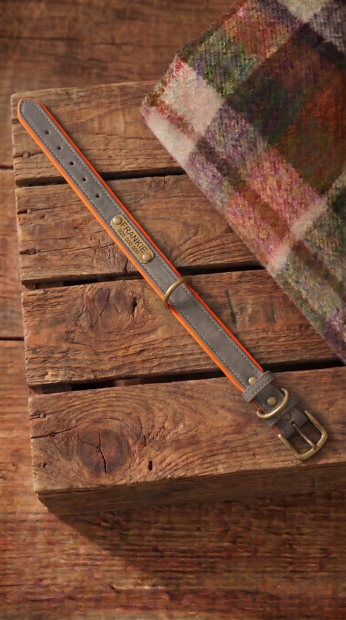 A leather strap with a gray center, orange stitching, and a brass buckle, laid diagonally on a rustic wooden block next to a folded plaid textile.